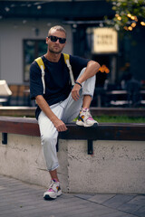 Young handsome bearded man in stylish black clothes looking through sunglasses, with a fashionable hairstyle, with backpack relaxing on the street. A handsome modern guy. Street style.
