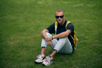 Young handsome bearded man in stylish black clothes looking through sunglasses, with a fashionable hairstyle, with backpack relaxing on the street. A handsome modern guy. Street style.
