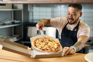 Pleasant young man in cook apron packing pizza into box