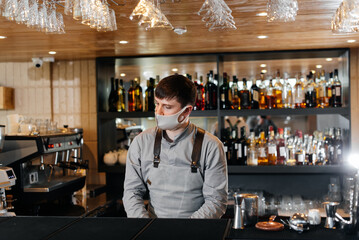 A stylish bartender in a mask and uniform during a pandemic is preparing cocktails at a party. The work of restaurants and cafes during the pandemic.