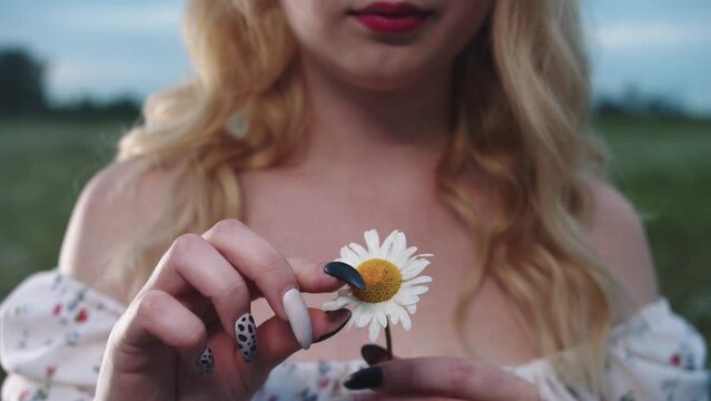 A young blonde woman tears off the petals of a daisy standing in a green field