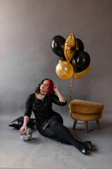 a girl with one half red hair the other black in a black tight dress with sequins for her birthday for a holiday in the studio on a gray background with golden black balloons with disco ball