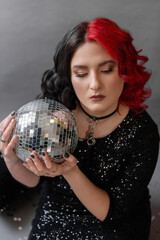 Naklejka premium a girl with one half red hair the other black in a black tight dress with sequins for her birthday for a holiday in the studio on a gray background with golden black balloons with disco ball