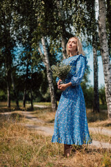 A young beautiful woman in a blue dress holds wild flowers in her hands