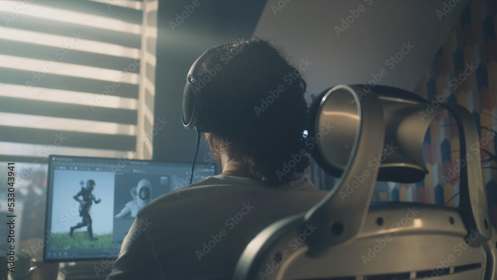 Poster back view of man in headphones sitting at the table and creating video game hero and 3d animation in