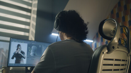 Back view of man in headphones sitting at the table and creating video game hero and 3D animation...