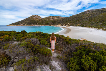 Fototapeta na wymiar a girl in a long colourful dress walks along a paradise beach with white sand and huge rocks and green mountains in the background; two people's bay, western australia