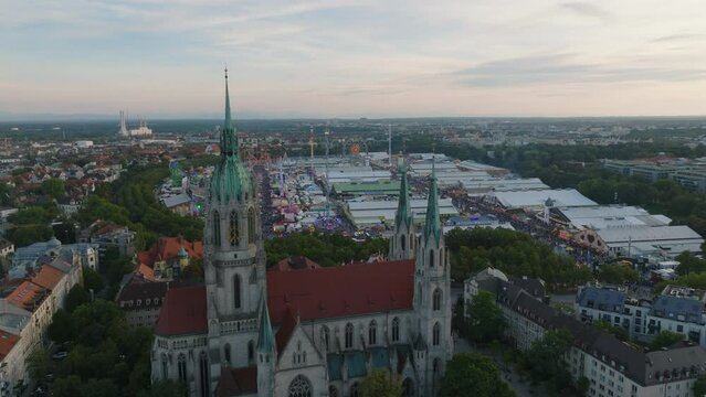 Aerial panoramic view of city at twilight. Fly over large gothic church and revealing Theresienwiese while visitors enjoying beer and atmosphere of Oktoberfest. Munich, Germany