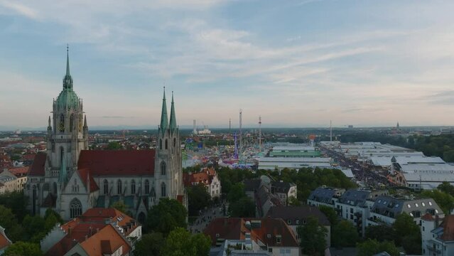Aerial ascending footage of gothic church in city at twilight. Revealing large beer tents and colourfully blinking attractions on Theresienwiese, Oktoberfest site. Munich, Germany
