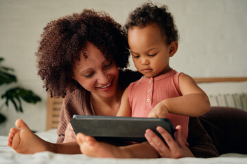 Happy biracial mother and daughter in casual clothing using digital tablet on bed at home