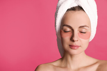 Beautiful young woman with under eye patches and hair wrapped in towel on pink background, space for text