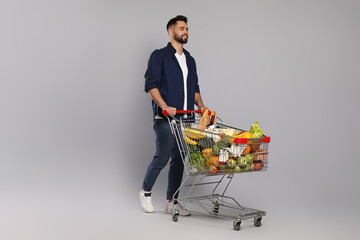 Happy man with shopping cart full of groceries on light grey background