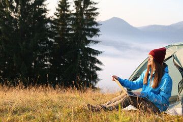 Woman working on laptop near camping tent outdoors surrounded by beautiful nature