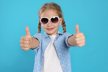 Girl in stylish sunglasses showing thumbs up on light blue background