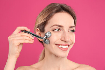 Young woman using metal face roller on pink background