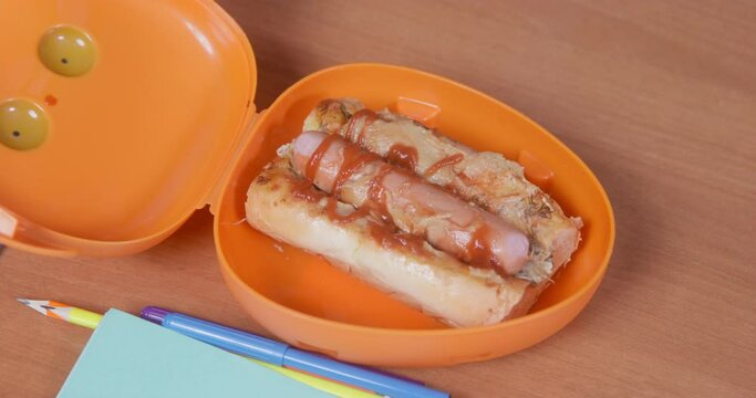 Dog In A Blue T-shirt Moves To Someone Open Lunchbox With A Paw, In Which There Is A Delicious Hot Dog With Sauce, Top View. Surface Of Table Is Littered With Stationery