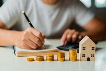 Businessman using calculator to calculate and taking notes and Stacking coins, wooden house on table. Saving money,accounting, investment, budgeting, and financial planning concept.