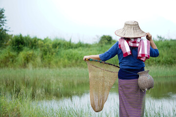 Back view of Asian woman farmer wears hat and traditional costume, stands at the pond and holds...