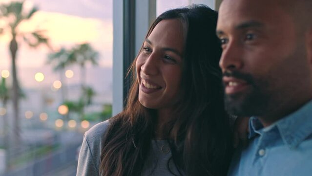 Happy Couple On Vacation, Watch The Sunset As Woman Points Outside And Man Gives Her A Kiss Enjoying Their Holiday. People Looking At View Together, In Their Luxury Modern Home At Twilight