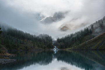 Mountain creek flows from forest hills into glacial lake. Snow rock mountains in fog clearance. Small river and coniferous trees reflected in calm alpine lake. Tranquil misty scenery at early morning.