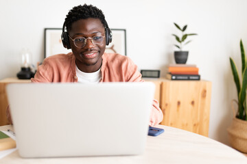Front view of young African American man wearing headphones working, studying at home using laptop. Copy space.