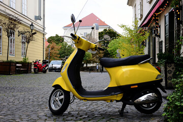 Trendy yellow Italian made scooter in old street. cobblestone road pavement. green trees and decorative planters. urban setting. white windows and stucco house facade. selective focus. soft background