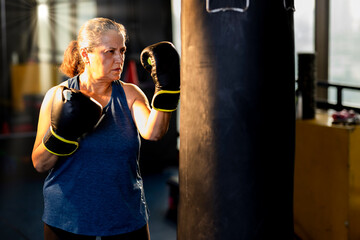Aggressive old woman in sportswear training with boxing bag in gym
 - Powered by Adobe