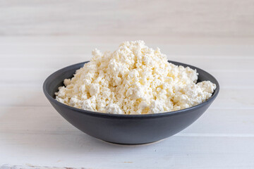 Cottage cheese in dark bowl on white wooden background.