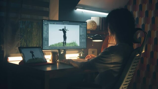 Guy sitting at the table at home at night and making 3D animation for video game usign modern personal computer and digital tablet