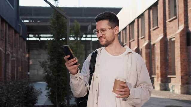 Positive happy Man holding smartphone, chatting in social media outdoor. Good looking male with coffee to go messaging on mobile phone on urban street. Businessman typing a message outside.