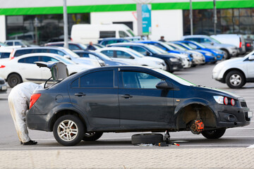 A car with a wheel removed is on a jack in a supermarket parking lot. The male driver takes a tool out of the trunk to fix a flat tire.