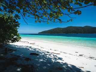 Scenic view of Koh Ra Wi Island white sand beach, clear turquoise sea against summer blue sky with Koh Adang Island background and tree shade foreground. Near Koh Lipe Island, Satun, Thailand.