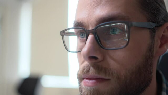 Close up macro shot of young Caucasian IT specialist sitting in office coding on computer. Program code reflects in glasses of guy programmer. Male face in eyeglasses in cabinet working on computer