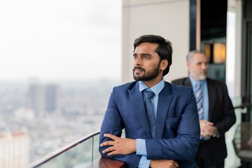 Portrait of a mature asian businessman standing looking out the office balcony with another caucasian businessman at the background out of focus
