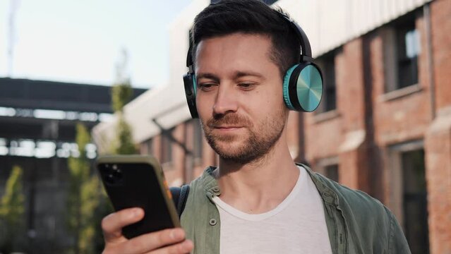 Handsome man in green white casual clothes wear wireless headphones listening to music using mobile phone app outdoors on an empty street, relaxing after work, male freelancer feel stress free relief.