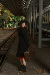 Portrait of middle-aged woman in coat and hat. Vintage lady with retro suitcase waiting train at railroad station