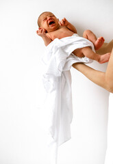 Young crying baby boy covered in white blanket in female hands on white wall background.