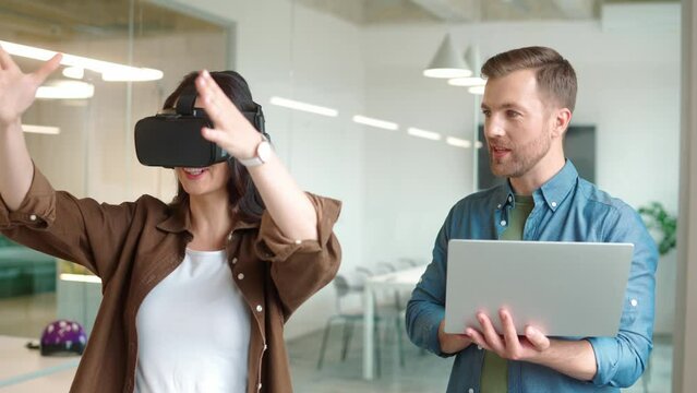Close up portrait of handsome young male manager holding laptop and speaking while woman wearing VR headset and moving hands in air. Virtual reality presentation. Office work. Futuristic technology