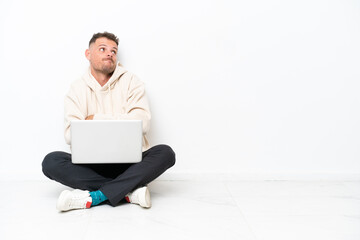 Young caucasian man with a laptop sitting on the floor isolated on white background making doubts gesture while lifting the shoulders