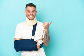 Young caucasian man wearing a sling  and neck lace isolated on blue background pointing to the side...