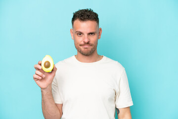Young caucasian man holding an avocado isolated on blue background with sad expression