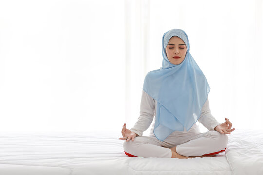 Young asian woman sitting on bed and enjoying meditation. Beautiful girl in muslim sleepwear with blue hijab practices yoga in bedroom with peace and calm. Healthy and lifestyle concept - Powered by Adobe