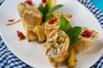 Simple homemade pancakes cut with banana, kiwi, strawberry jam on a white plate. Creamy curd filling. The food is decorated with mint. Rolled thin roll of pancake.