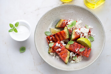 Plate with grilled watermelon batons, grated cheese and white yogurt, elevated view on a light-beige marble background, horizontal shot