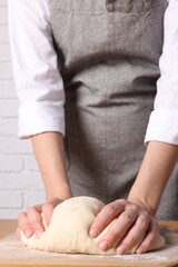 Woman kneading dough at wooden table near white brick wall, closeup