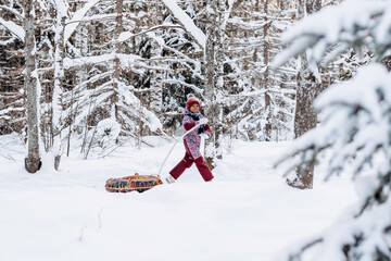 Happy little African-American girl in a red hat and jumpsuit rides on tubing in the winter park.Beautiful trees are covered with white snow.Winter fun,active lifestyle concept. - obrazy, fototapety, plakaty