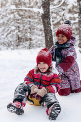 Happy Caucasian and African-American girls ride a saucer in the winter park.Beautiful trees are...