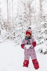 Fototapeta na wymiar Happy little African-American girl in red hat and jumpsuit walks in the winter forest and throws up snow.Beautiful trees are covered with white snow.Winter fun,active lifestyle concept.Selective focus