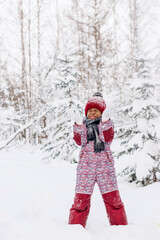 Happy little African-American girl in red hat and jumpsuit walks in the winter forest and throws up snow.Beautiful trees are covered with white snow.Winter fun,active lifestyle concept.Selective focus