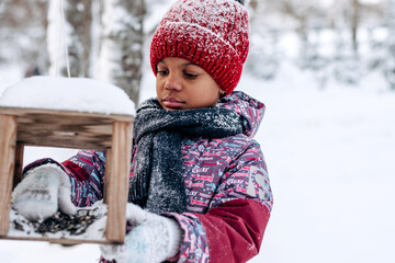Little African-American girl in a red hat and jumpsuit pours food into a bird feeder in a winter...